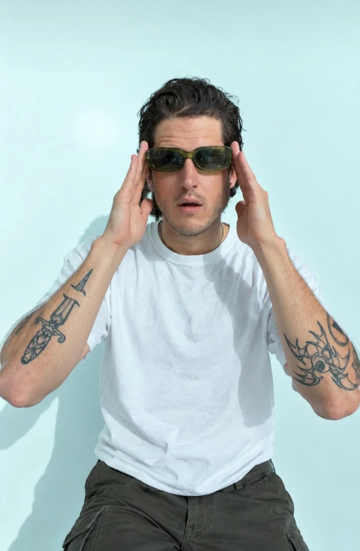 L.T. Drifta in green sunglasses and a white tee against a pale blue wall